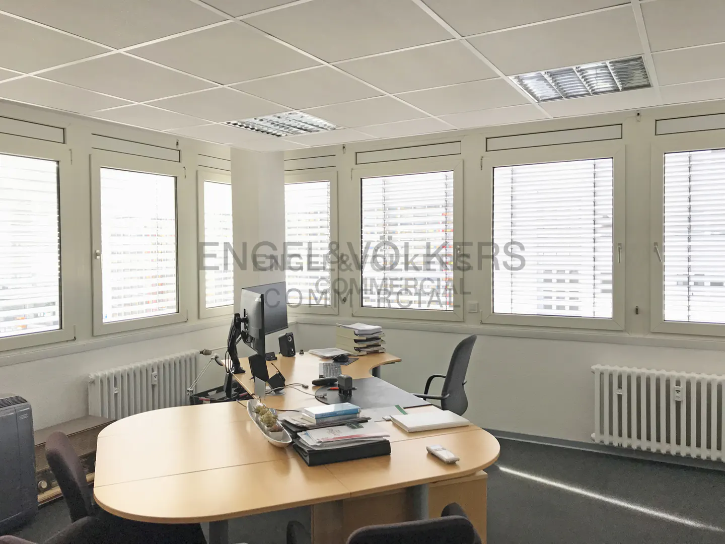 Bright office space with a large desk, computer, and multiple windows with blinds. Radiators are visible under the windows.