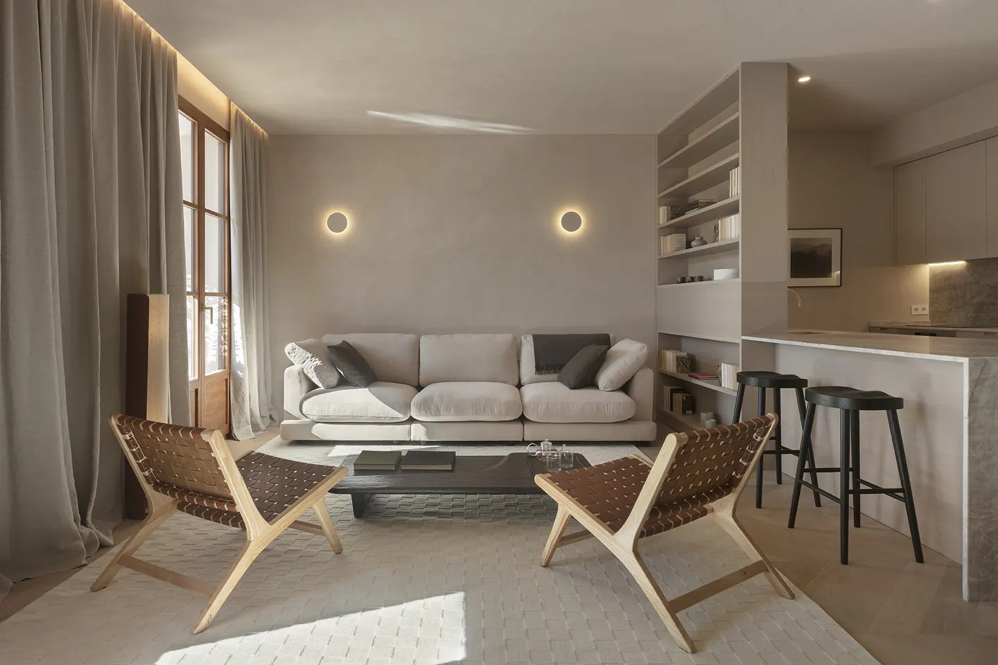 A modern living room with a beige sofa, two woven chairs, and a black coffee table on a patterned rug. A kitchen island with stools is visible in the background.