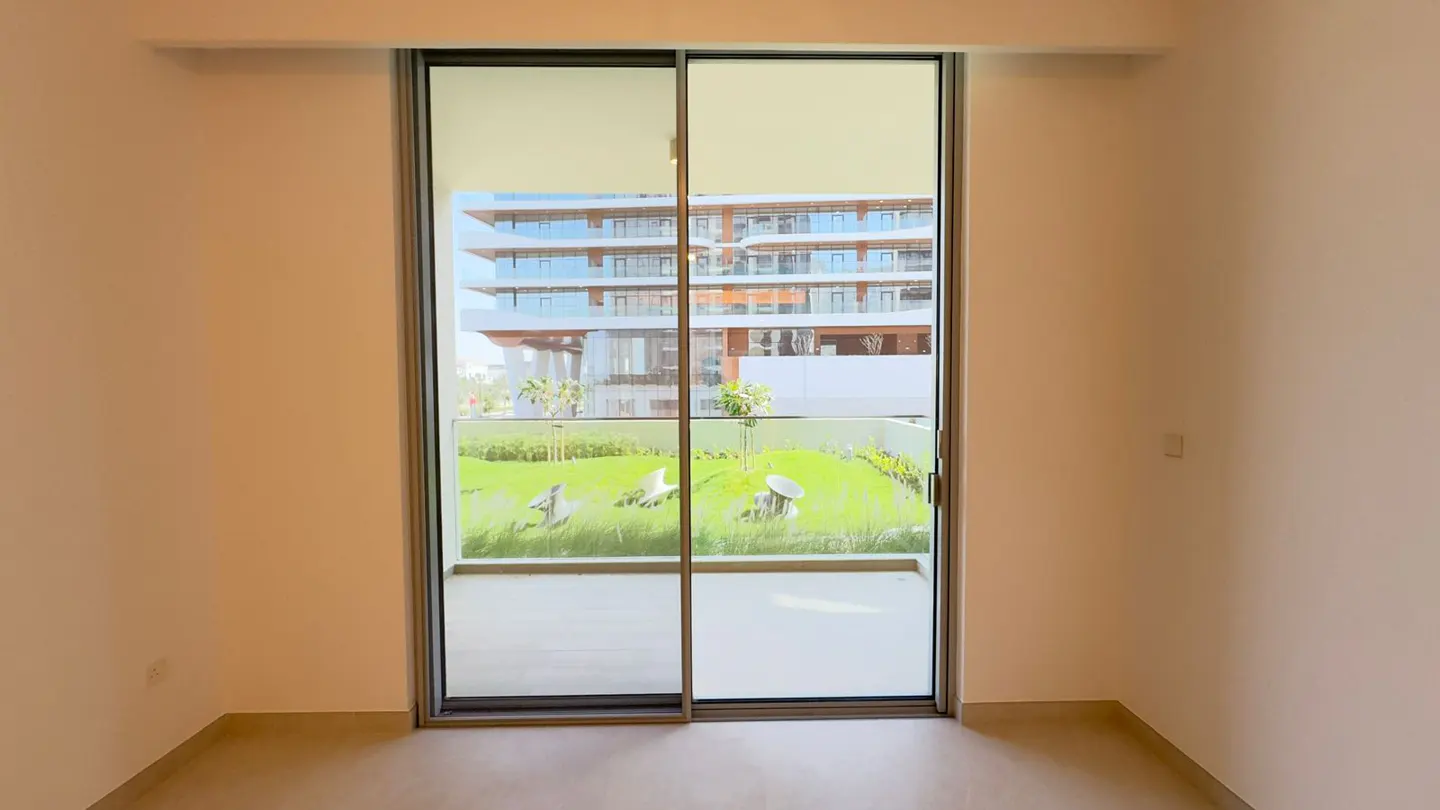 A room with beige walls and a sliding glass door leading to a balcony with a green lawn and a building in the background.