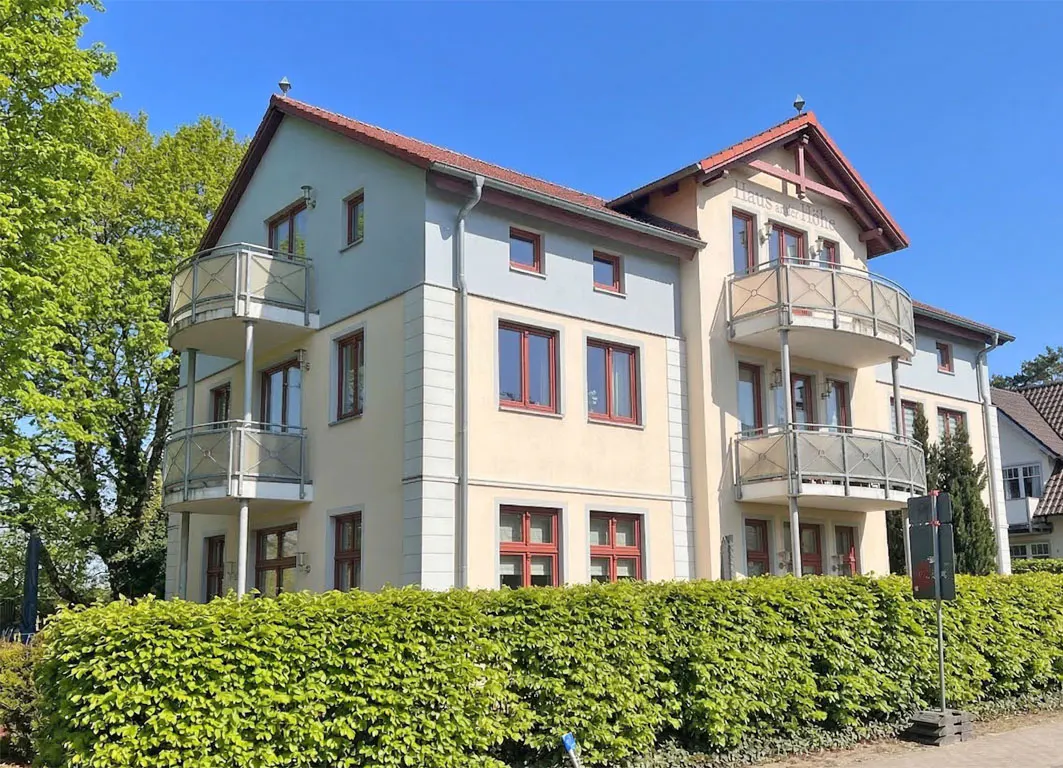 A three-story apartment building with balconies, cream and blue walls, and red-framed windows, behind a green hedge.