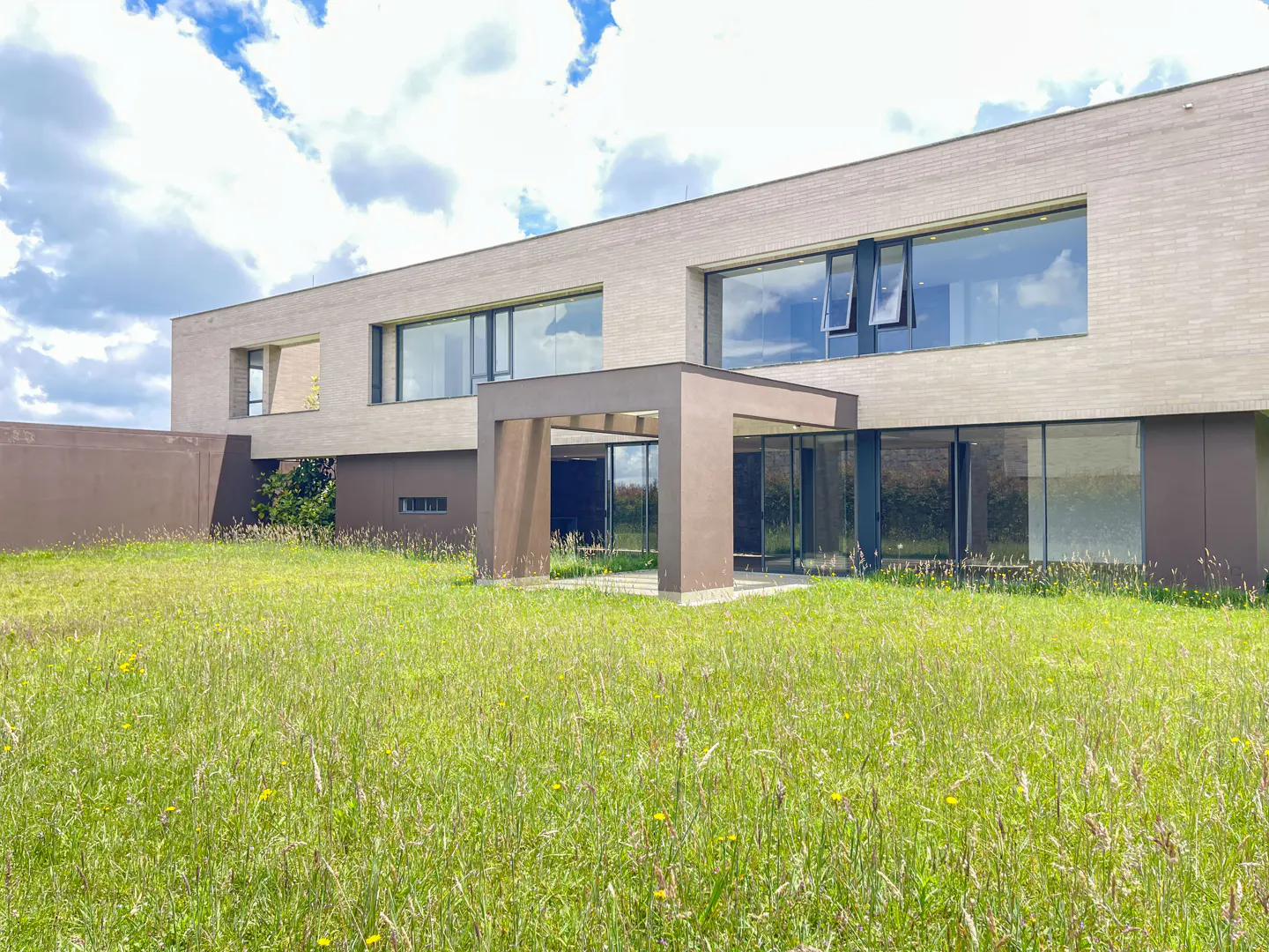 Modern beige brick house with large windows and a brown portico, surrounded by a green lawn under a cloudy sky.