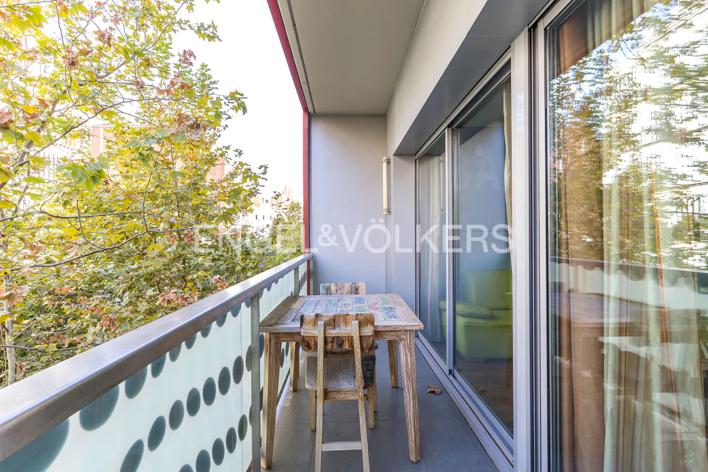 Balcony with a wooden table and chairs, overlooking trees. Sliding glass doors lead inside. Engel & Völkers logo visible.