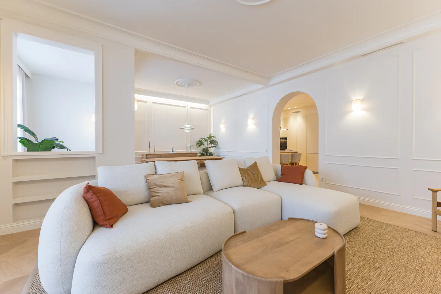 Bright living room with a white sectional sofa, rust and tan pillows, a wooden coffee table, and an arched doorway.