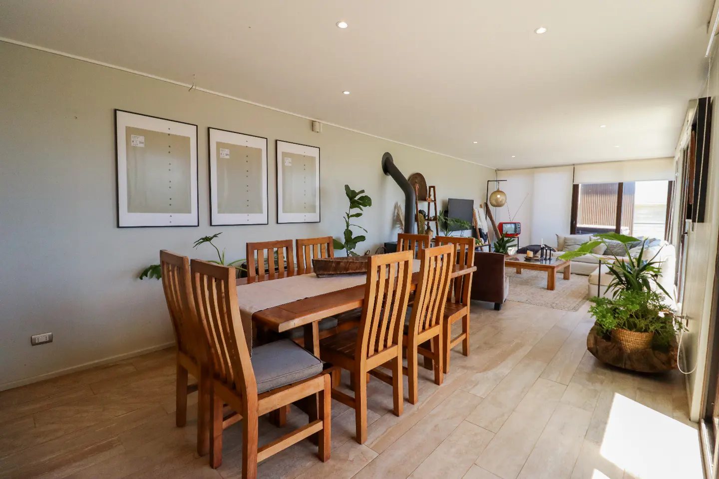 Open-plan dining and living area with a wooden table, six chairs, a wood-burning stove, and a bright, modern aesthetic.