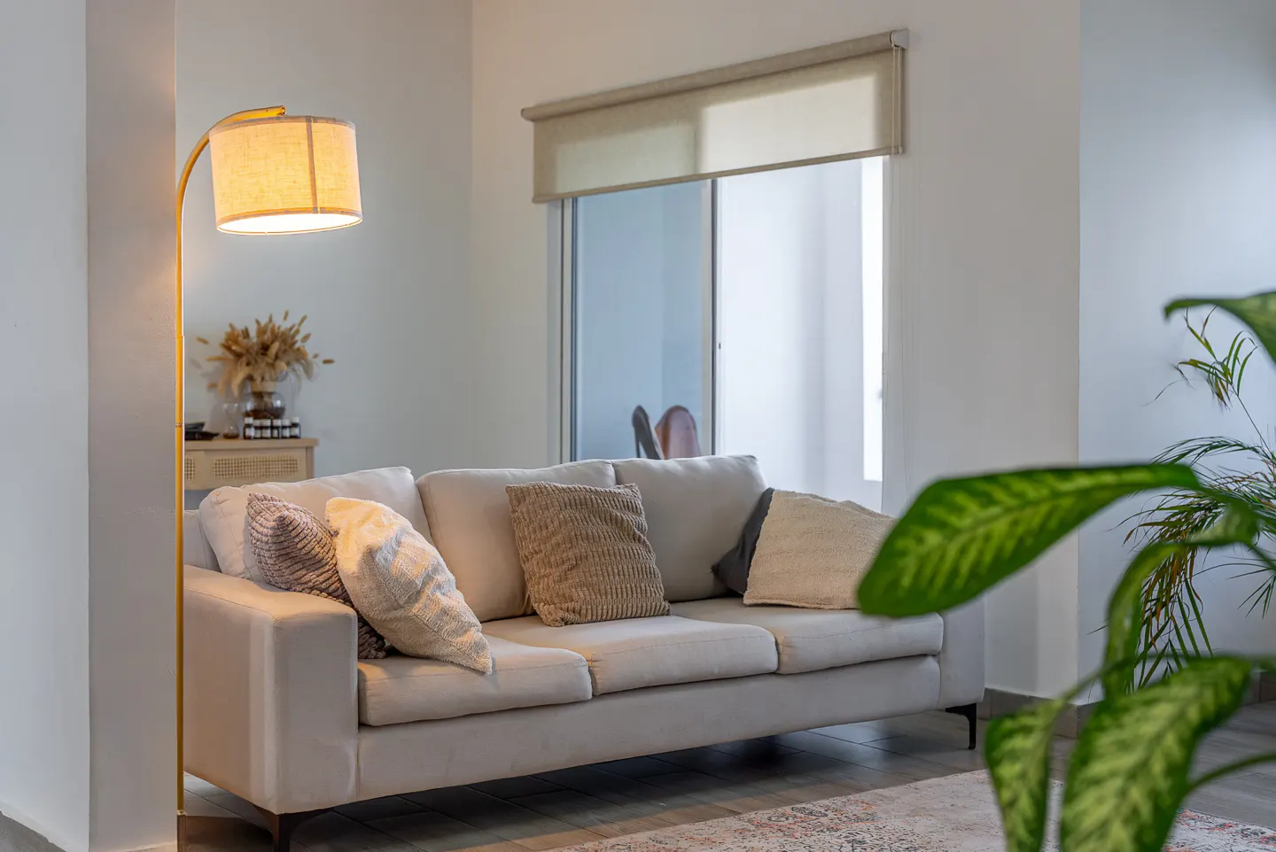 Bright living room with a beige sofa, decorative pillows, and a gold floor lamp. A large window with a shade is in the background.