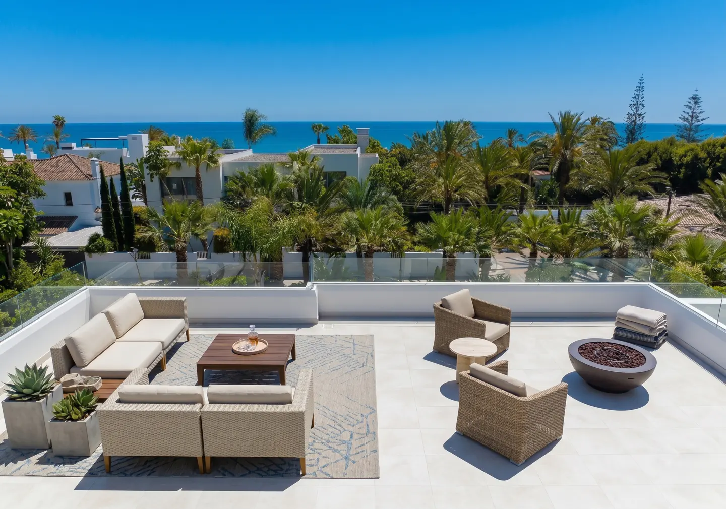 Rooftop patio with beige seating, a fire pit, and ocean views. Palm trees and white buildings are visible in the background under a clear blue sky.