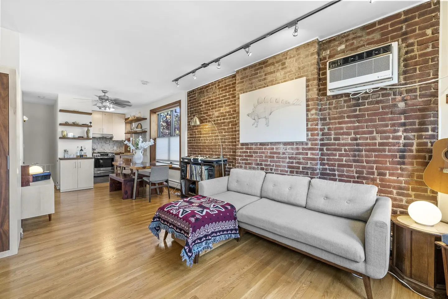 Living room with exposed brick wall, light gray sofa, and wooden floors. Kitchen and dining area visible in the background.