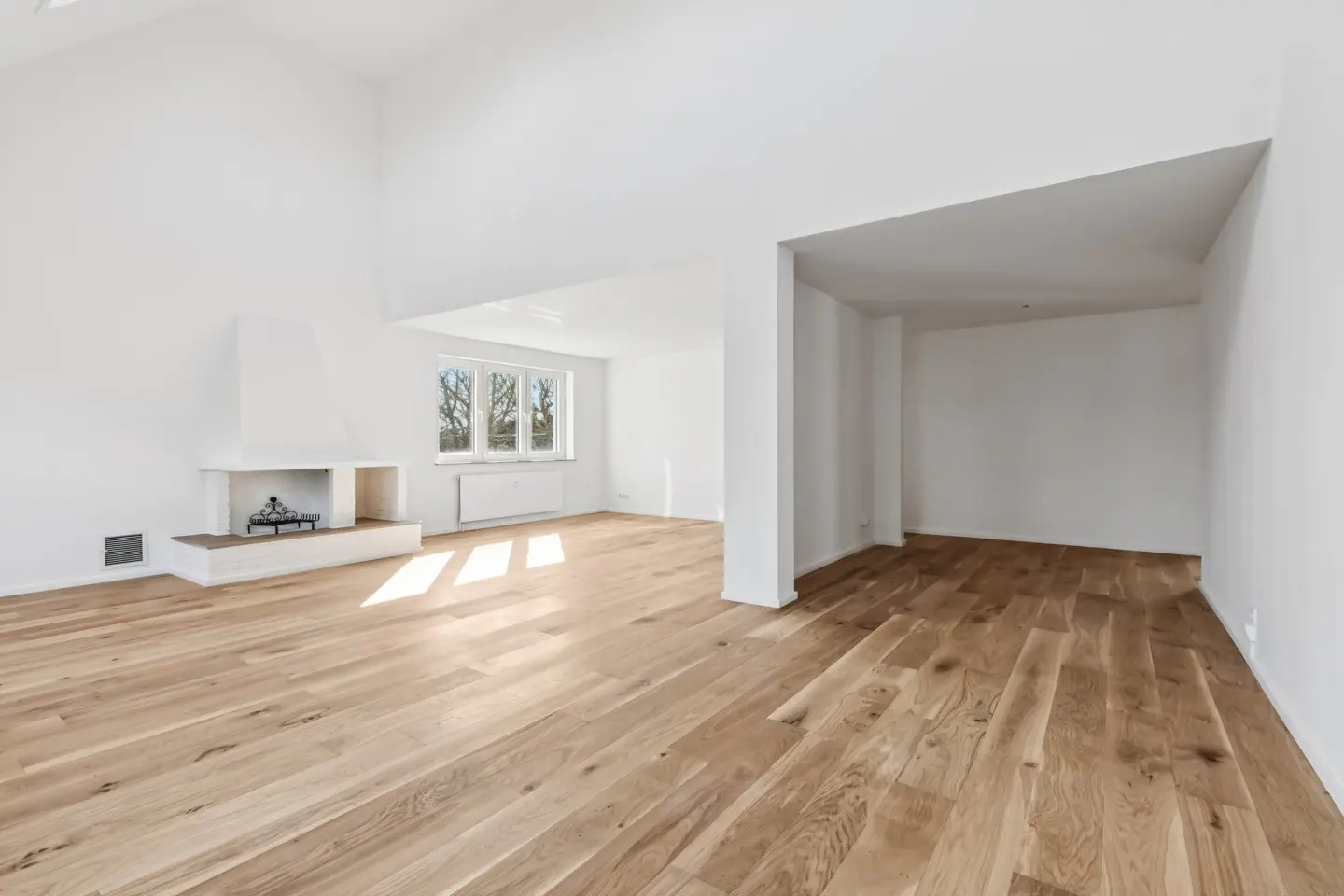 Bright, empty room with light wood floors, white walls, and a fireplace. A window lets in sunlight.