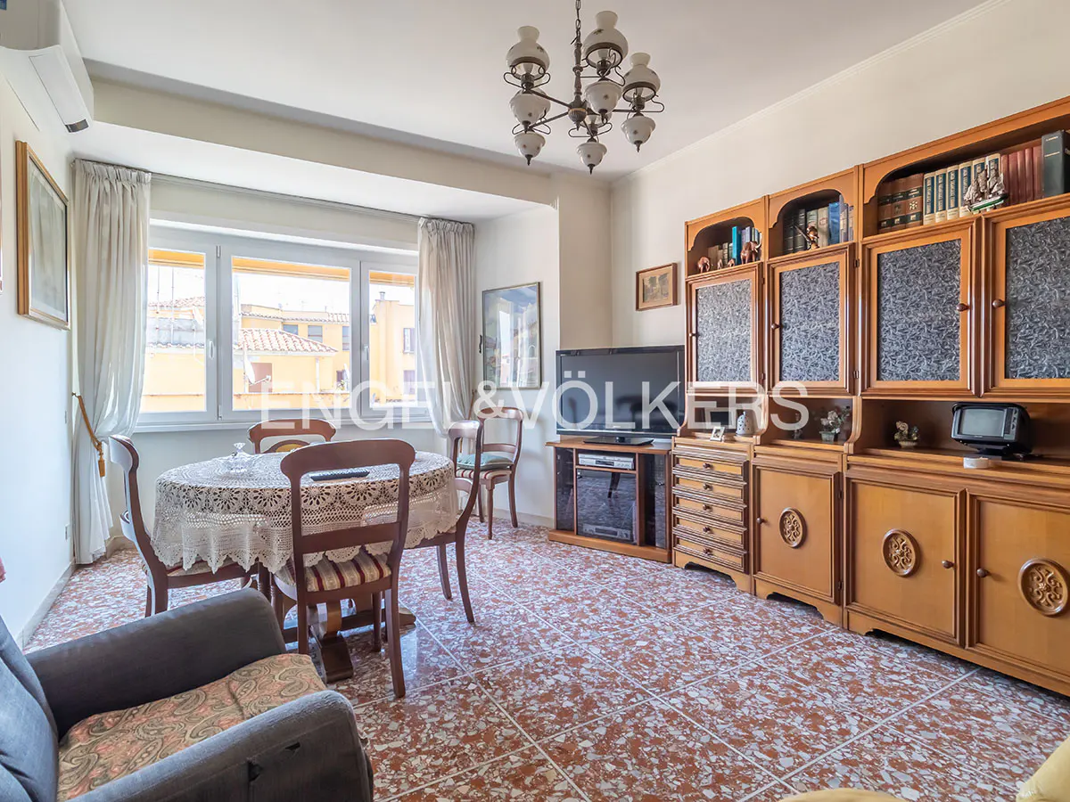 A living room with a round table, chairs, a wooden cabinet, and a TV. The room has a chandelier and a window with a view of buildings.