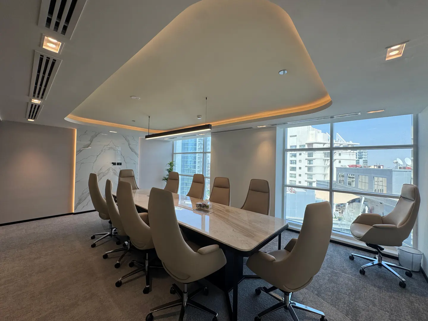 Modern conference room with a long marble table, beige ergonomic chairs, and large windows overlooking a city skyline.