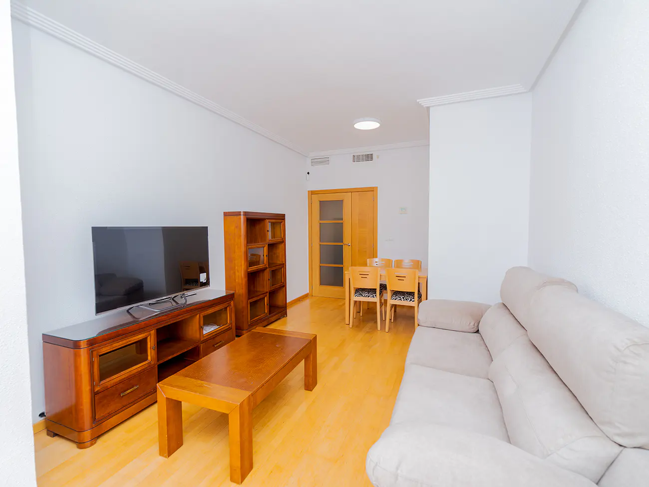 Bright living room with a beige sofa, wooden TV stand, flat screen TV, coffee table, bookcase, and dining table with four chairs.