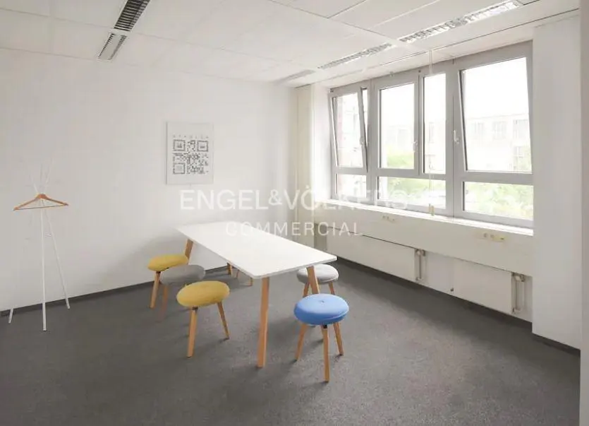 Bright office space with a white table, colorful stools, and large windows. A coat rack stands near a wall with a QR code poster.