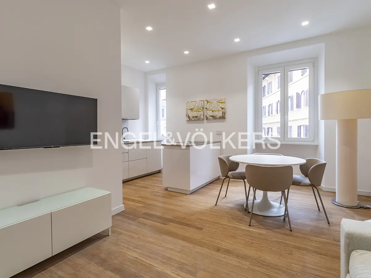 Bright, modern apartment with white walls, wood floors, and a flat-screen TV. A white table with four chairs sits near a window.