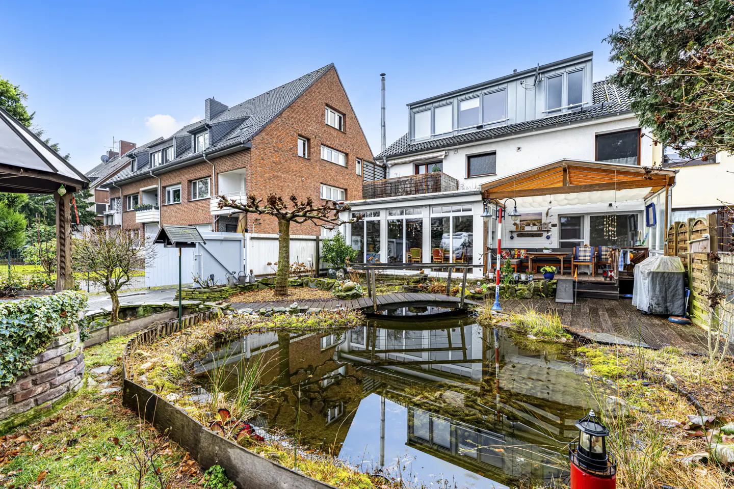 Backyard view with a pond reflecting the brick and white houses. A small wooden bridge crosses the pond. A deck and patio are visible.