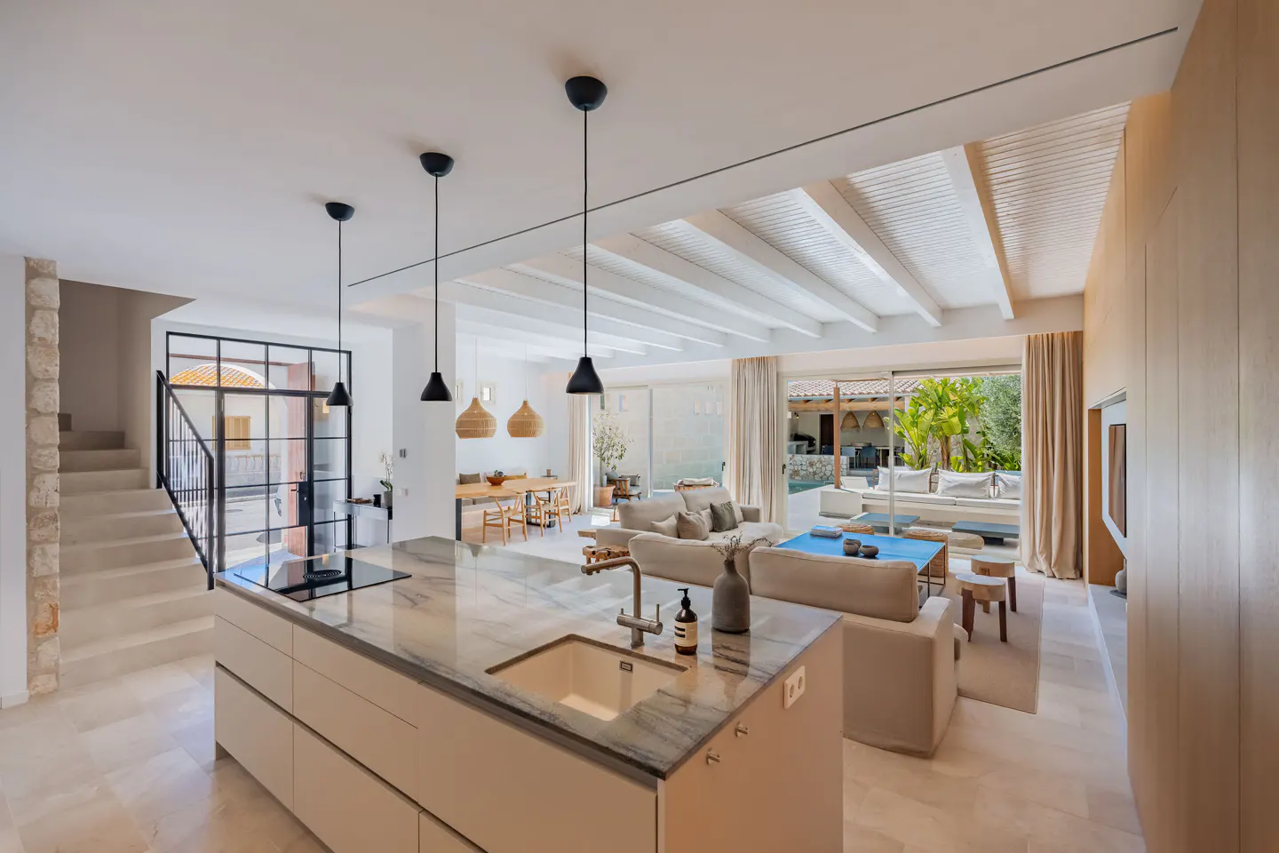 Open-concept living space with kitchen island, dining table, and living room. Neutral tones, white ceiling beams, and outdoor view.