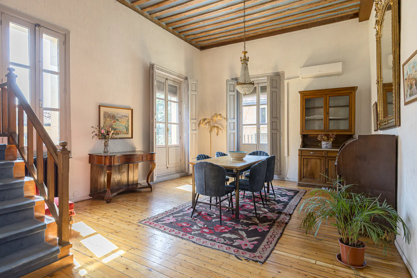 A dining room with wood floors, a table with blue chairs, and a patterned rug. A staircase is on the left, and a cabinet is on the right.