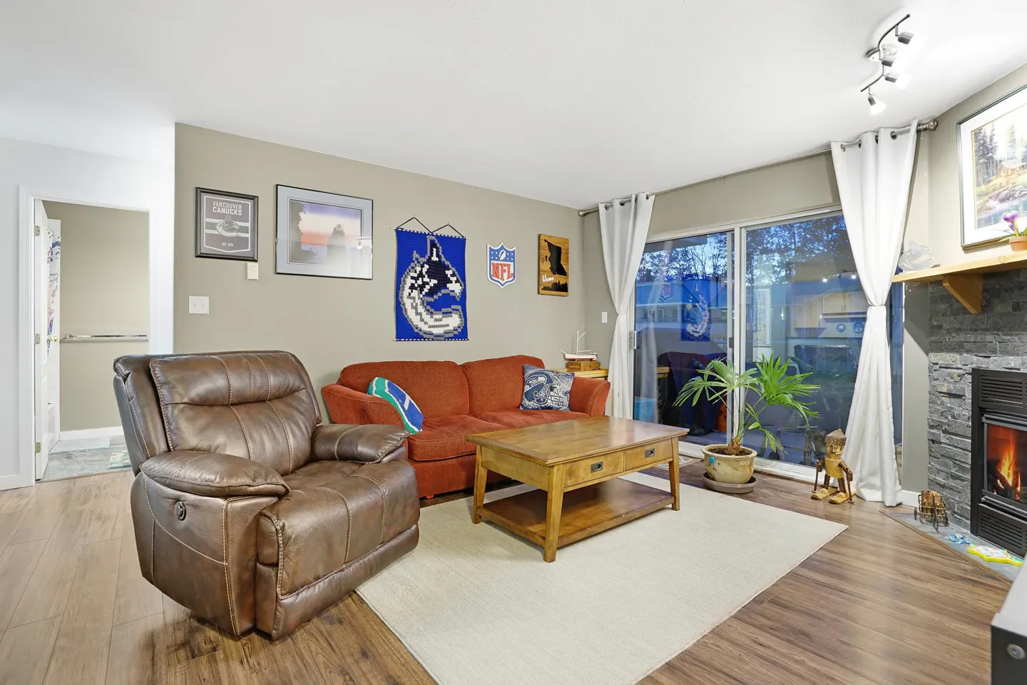 Living room with brown leather recliner, orange sofa, wood coffee table on a white rug. Sliding glass doors with white curtains lead to a patio. Fireplace on the right.