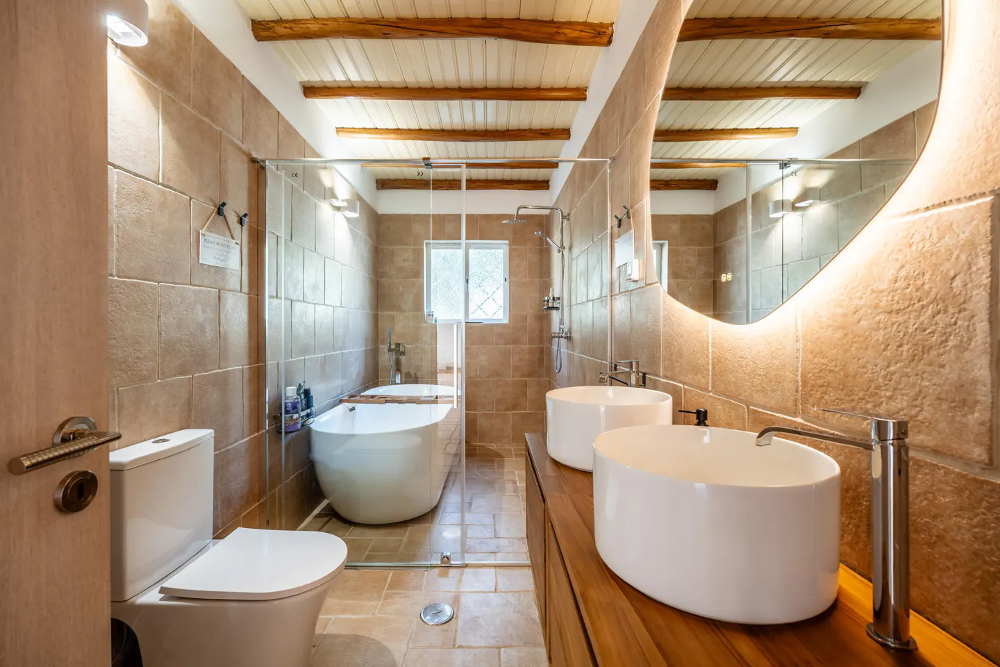 Bathroom with tan tile walls, wood ceiling beams, and a glass-enclosed tub. Two white vessel sinks sit on a wood vanity.