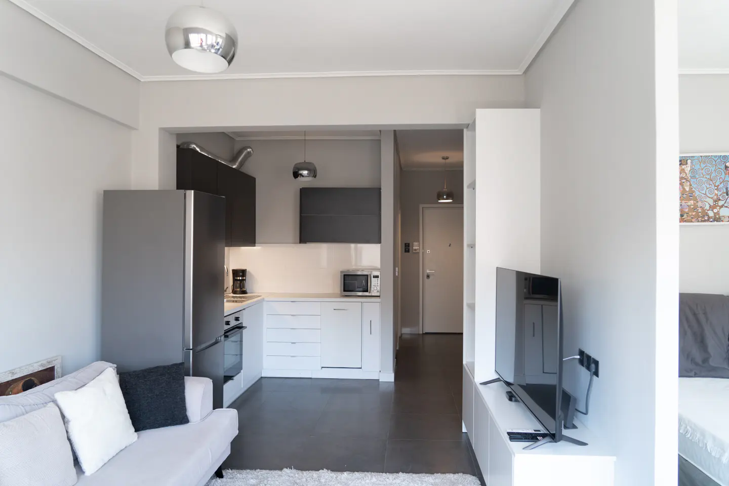 Modern open-plan apartment with a grey sofa, white kitchen cabinets, stainless steel fridge, and a TV on a white stand.