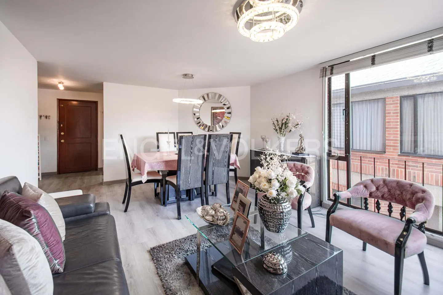 Bright living room with a dining table, gray sofa, glass coffee table, and pink chairs near a window.