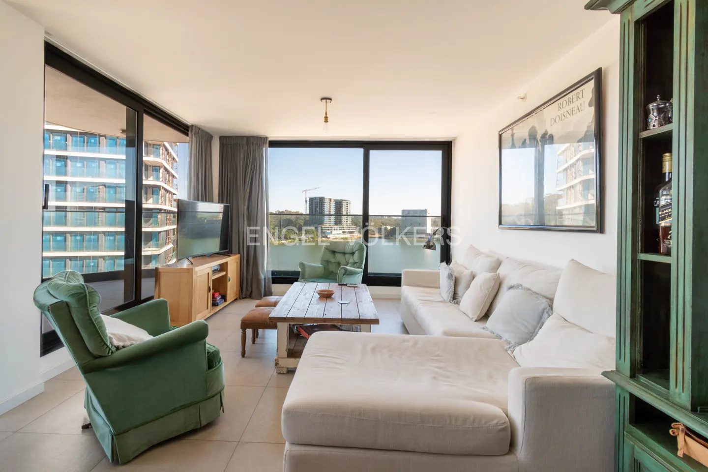 Bright living room with a white sectional sofa, green armchairs, and a wooden coffee table. Large windows offer city views.