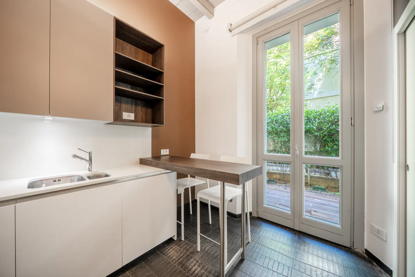 A modern kitchen with white cabinets, a stainless steel sink, and a dark wood countertop with two white stools. A window overlooks a green garden.