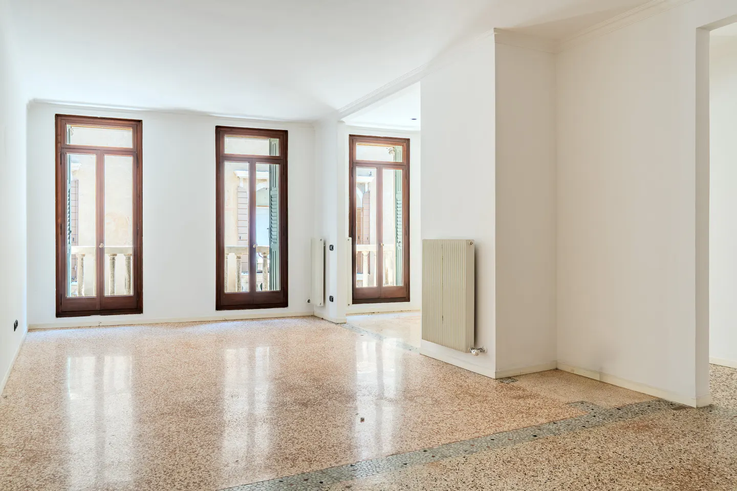 Empty room with white walls, polished terrazzo floors, and three tall wooden-framed glass doors leading to a balcony.