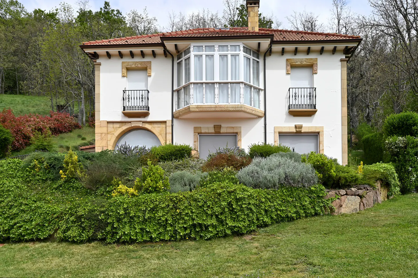 Two-story white house with a red tile roof, stone accents, and a bay window, surrounded by lush green landscaping.