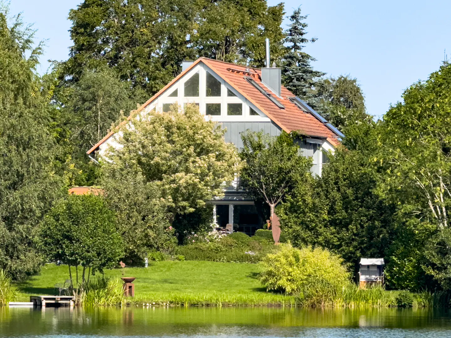 A modern house with a red roof is surrounded by lush green trees and a calm lake in the foreground.