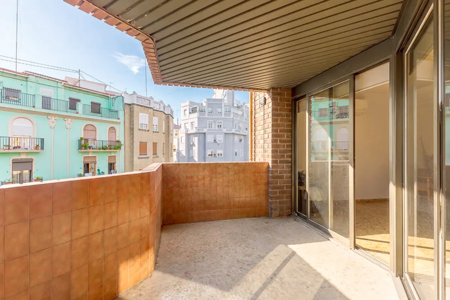 A balcony with brown tiled walls and a concrete floor, overlooking a city street with colorful apartment buildings.