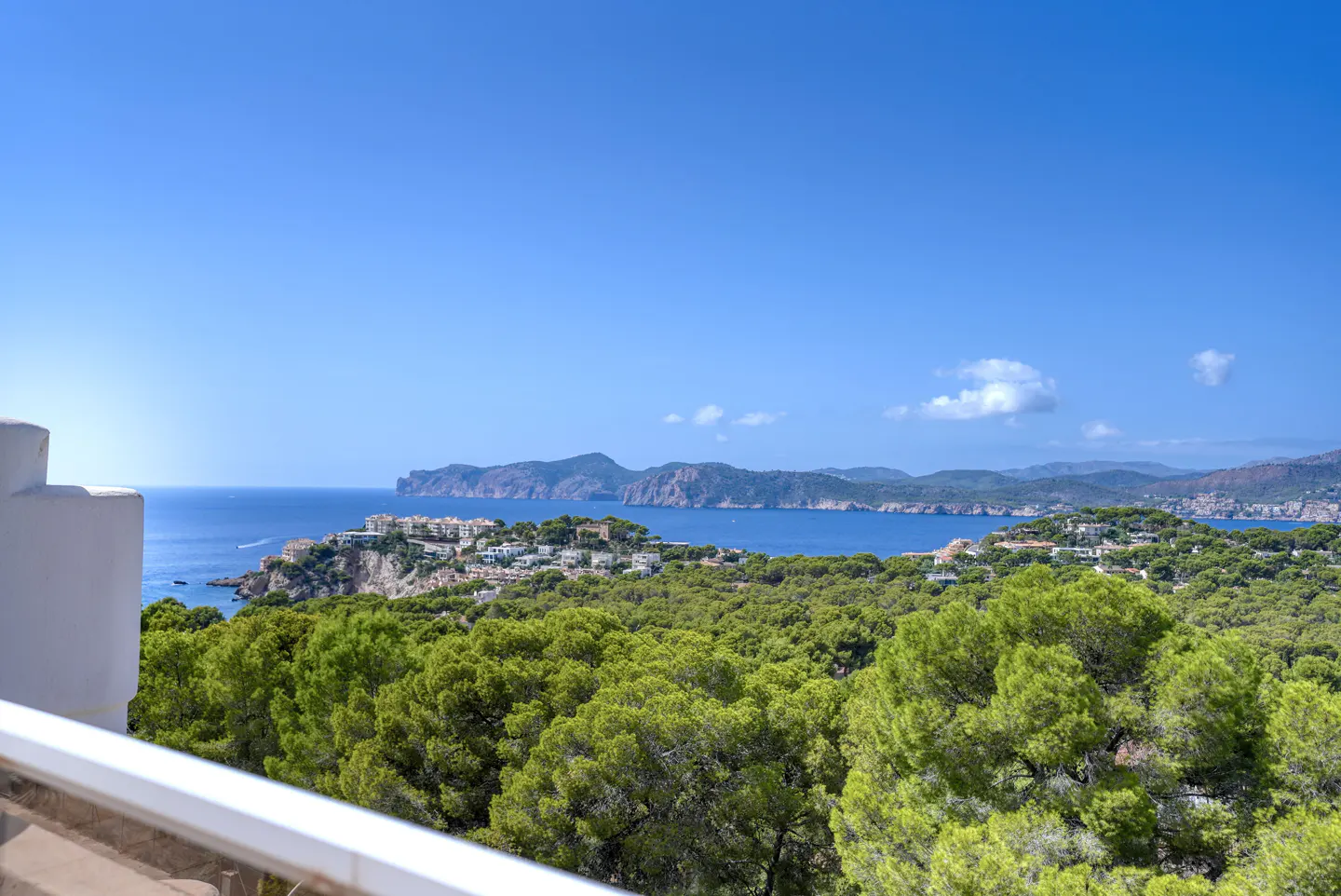 View from a balcony overlooking a lush green forest, blue ocean, and distant mountains under a clear blue sky.