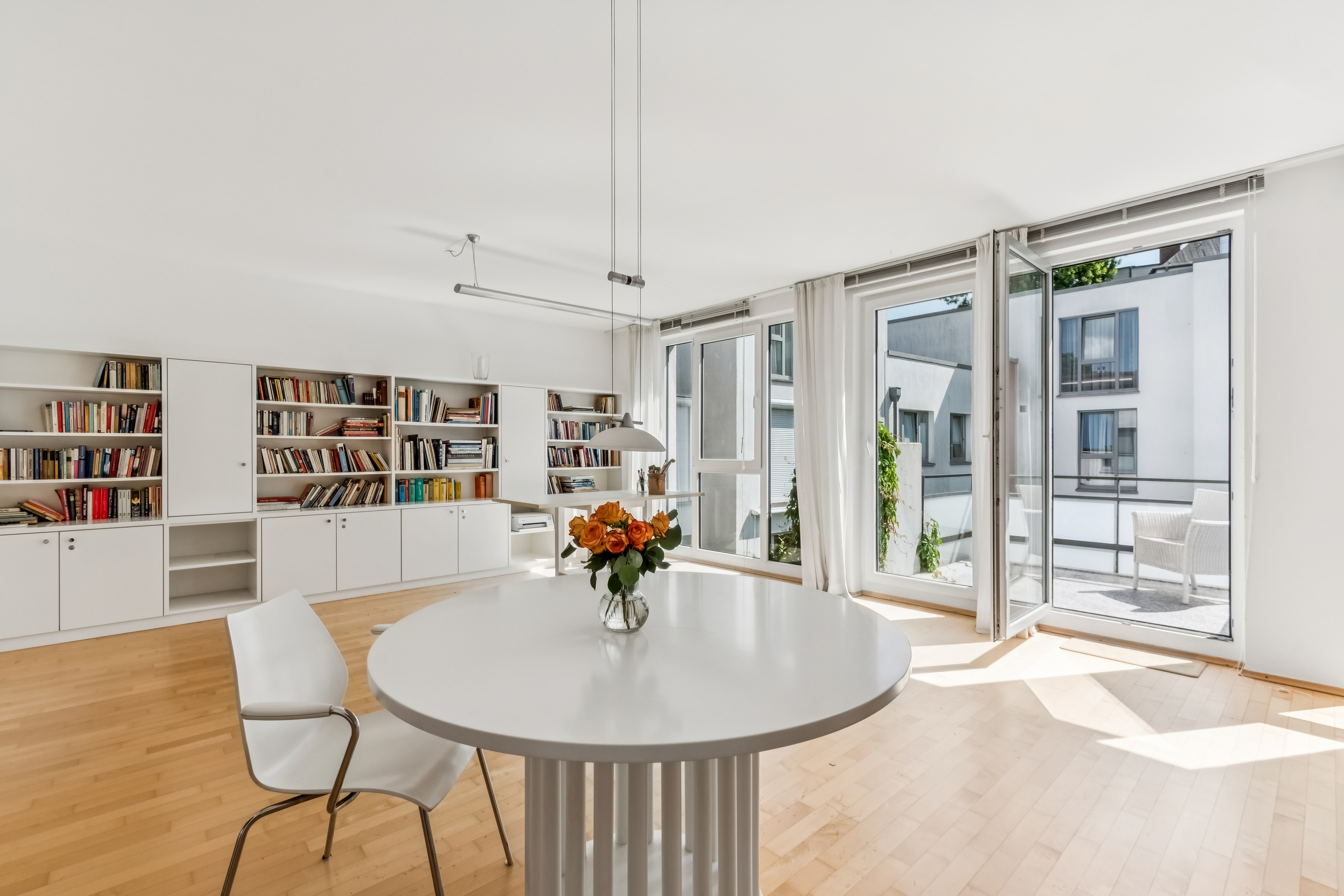 Bright living room with a white round table, orange flowers, a white chair, and a large wall-to-wall bookshelf.