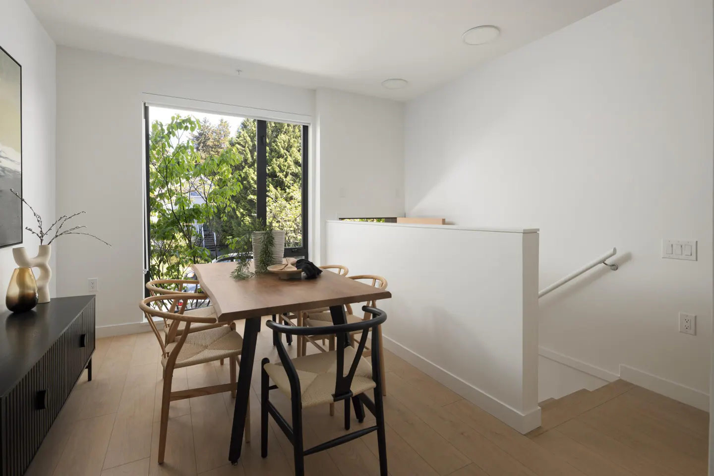 Bright dining area with a wooden table, woven chairs, and a black sideboard. A large window overlooks green trees. White walls and light wood floors.