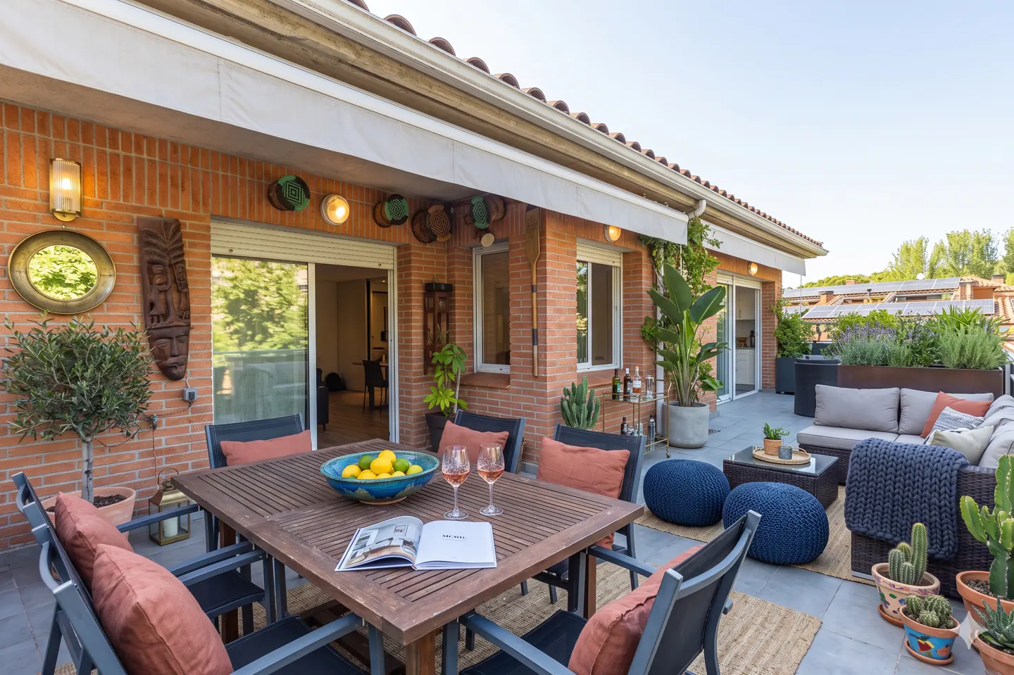 Spacious brick terrace with a wooden dining set, lounge area, potted cacti, and decorative wall art under a sun awning.