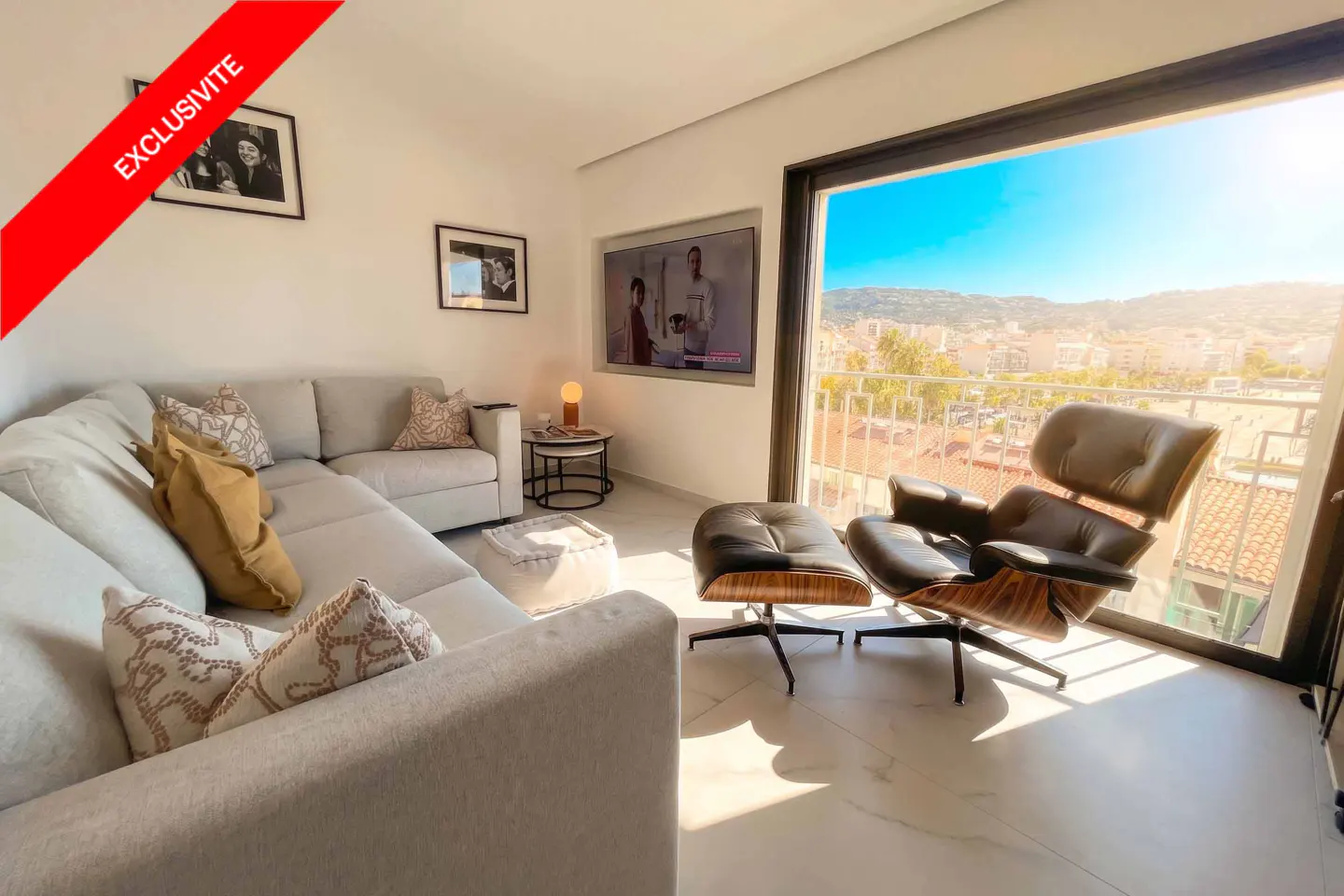 Bright living room with a beige sectional sofa, leather chair, and a large window with a city view. "Exclusivite" banner on the top left.