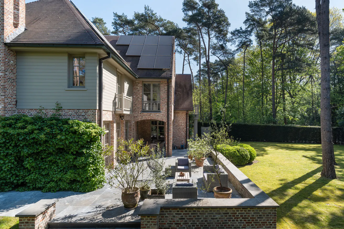Brick house with solar panels, a stone patio with outdoor furniture, and a green lawn surrounded by trees.