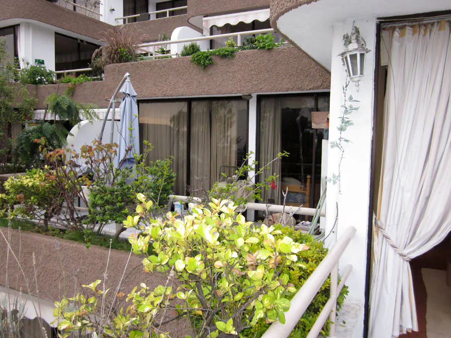 Exterior view of a multi-story building with balconies, plants, and a blue umbrella. A white lamp is on the wall.
