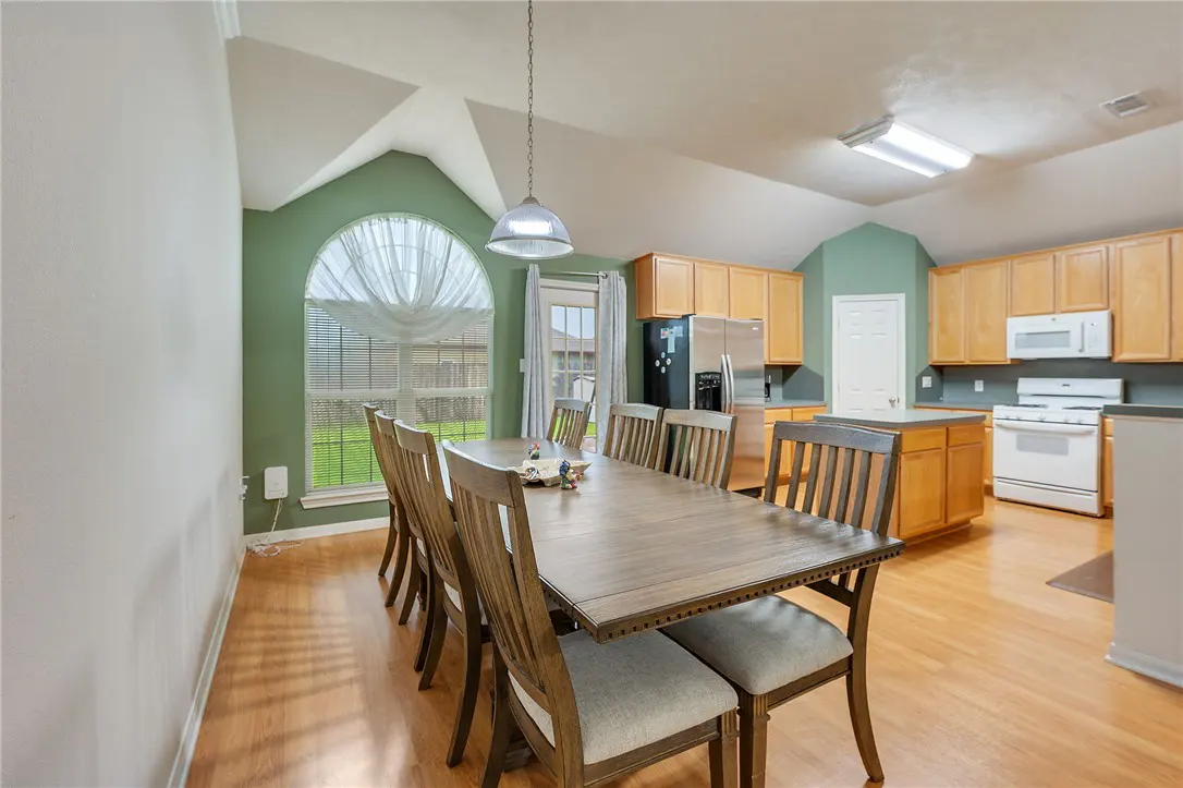 A dining room with a long wooden table and chairs, hardwood floors, and a kitchen in the background.