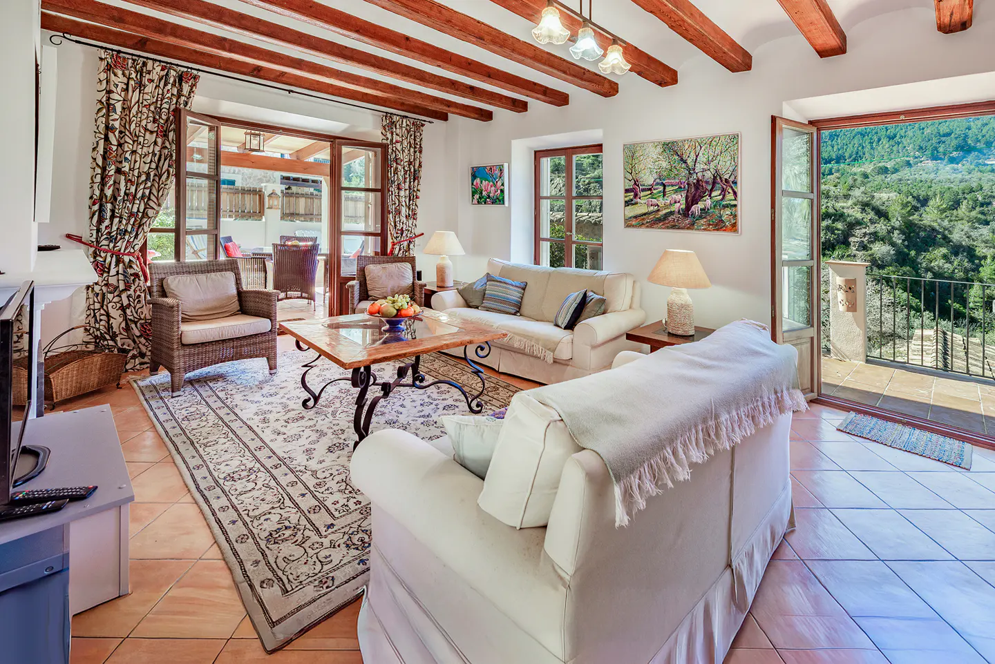 Bright living room with white sofas, wood beams, and open doors to a balcony with a view of green hills.