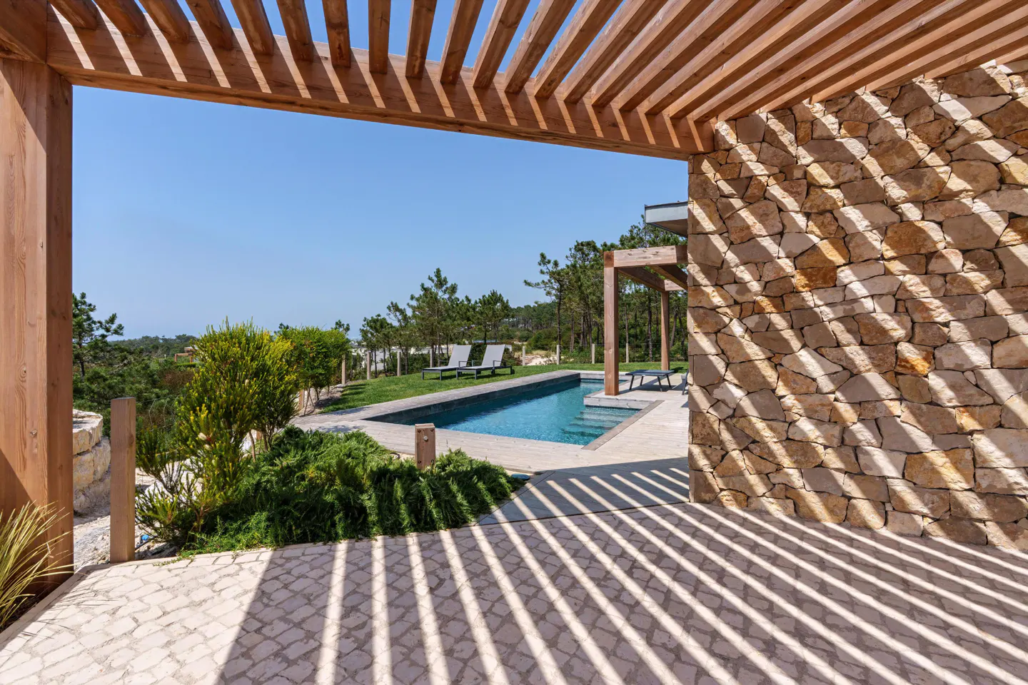 View of a blue pool and lounge chairs through a wooden pergola and stone wall on a sunny day.
