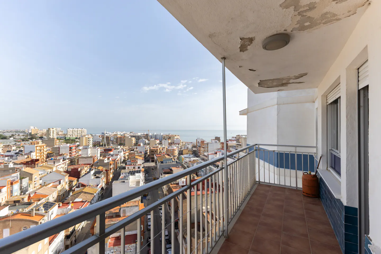 Balcony view of a city with the ocean in the background. The balcony has a metal railing and brown tile floor.