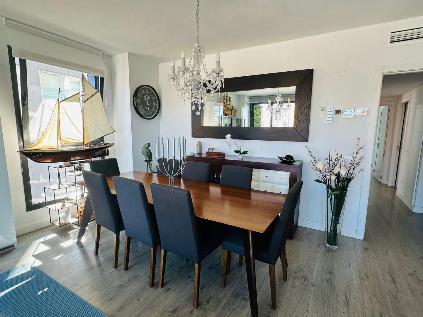 Bright dining room with wood table, blue chairs, chandelier, and a large mirror over a sideboard. A model sailboat sits by the window.
