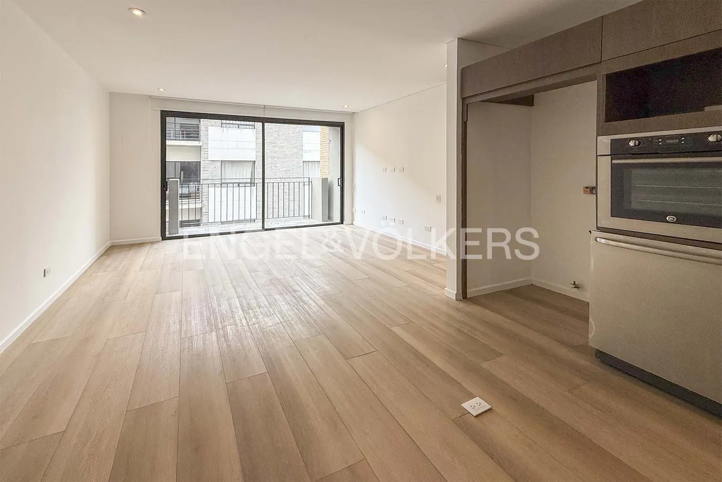 Bright, modern apartment interior with light wood floors, white walls, and a sliding glass door to a balcony. Stainless steel oven and cabinets are visible.