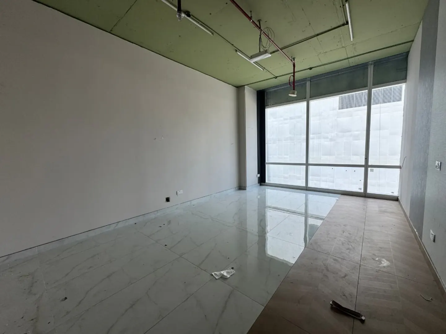 Unfurnished room with white walls, a green ceiling, and large windows. The floor is tiled in white marble. Overhead lighting and red pipes are visible.
