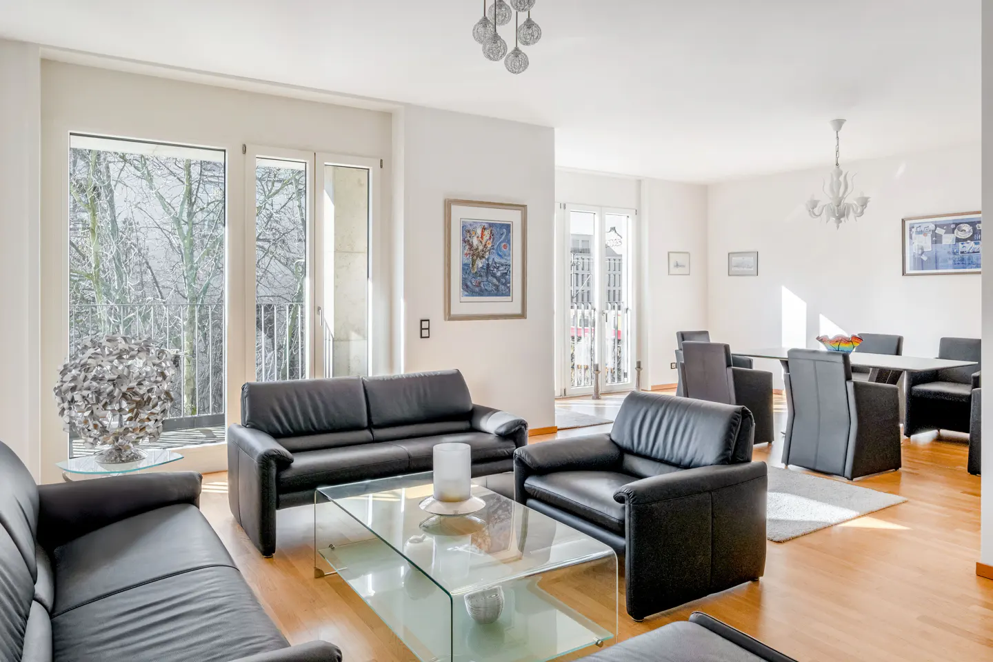 Bright living room with black leather sofas, glass tables, and wood floors. Dining area with table and chairs in the background.