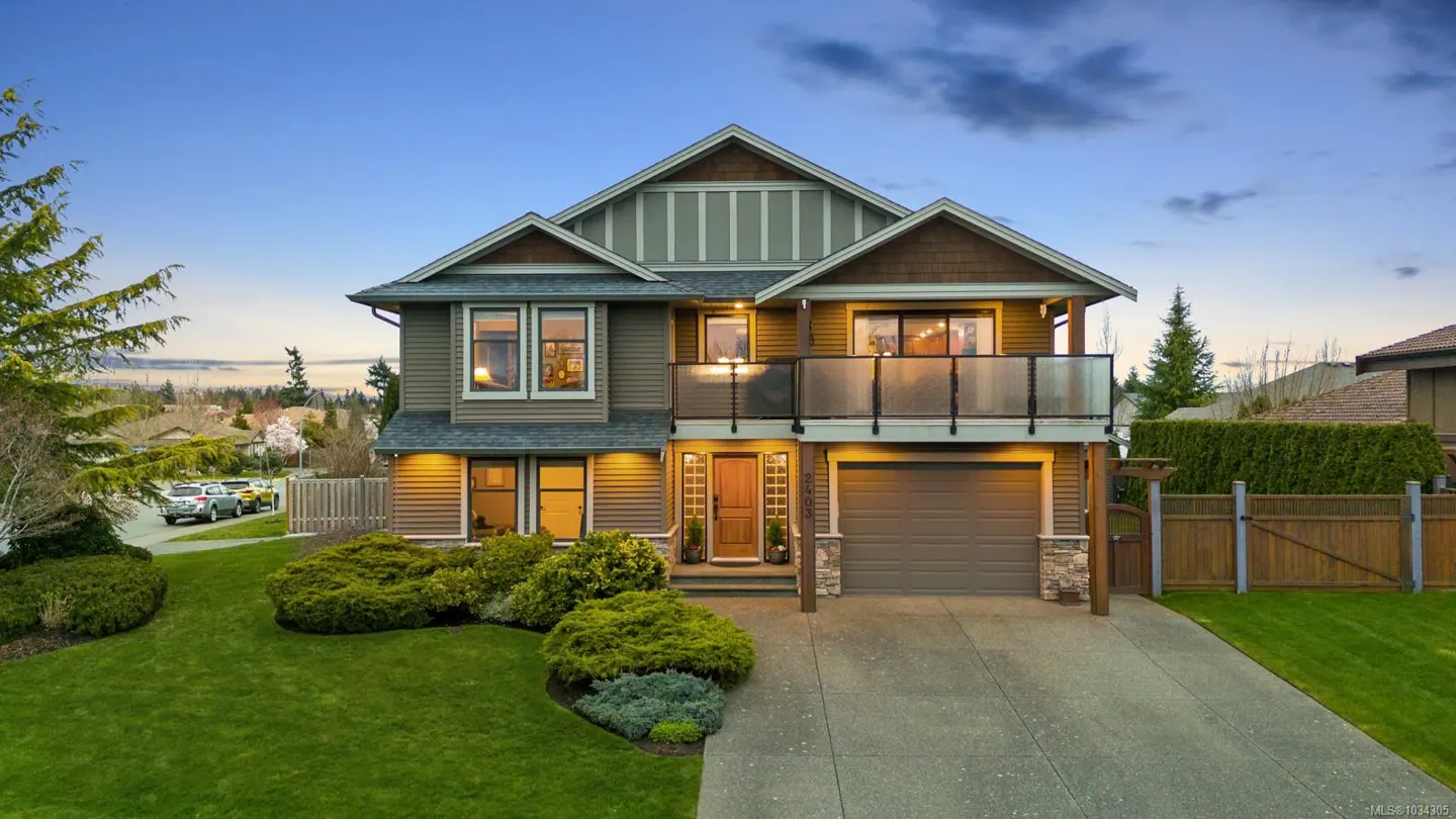Two-story house with brown siding, a balcony, and a garage. Green lawn and bushes in front. Blue sky in the background.