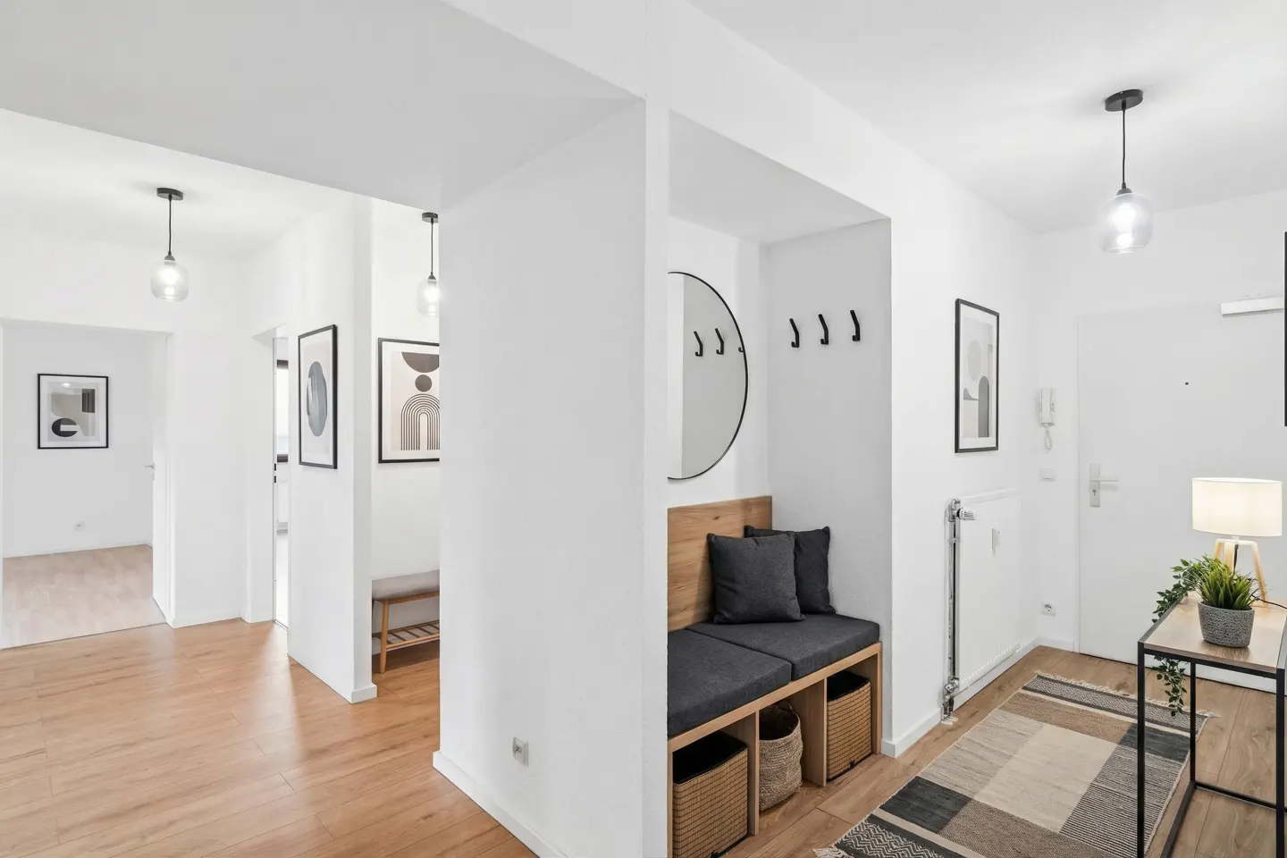 Bright, modern apartment entryway with white walls, wood floors, and a built-in bench with storage. A round mirror and coat hooks are above the bench.