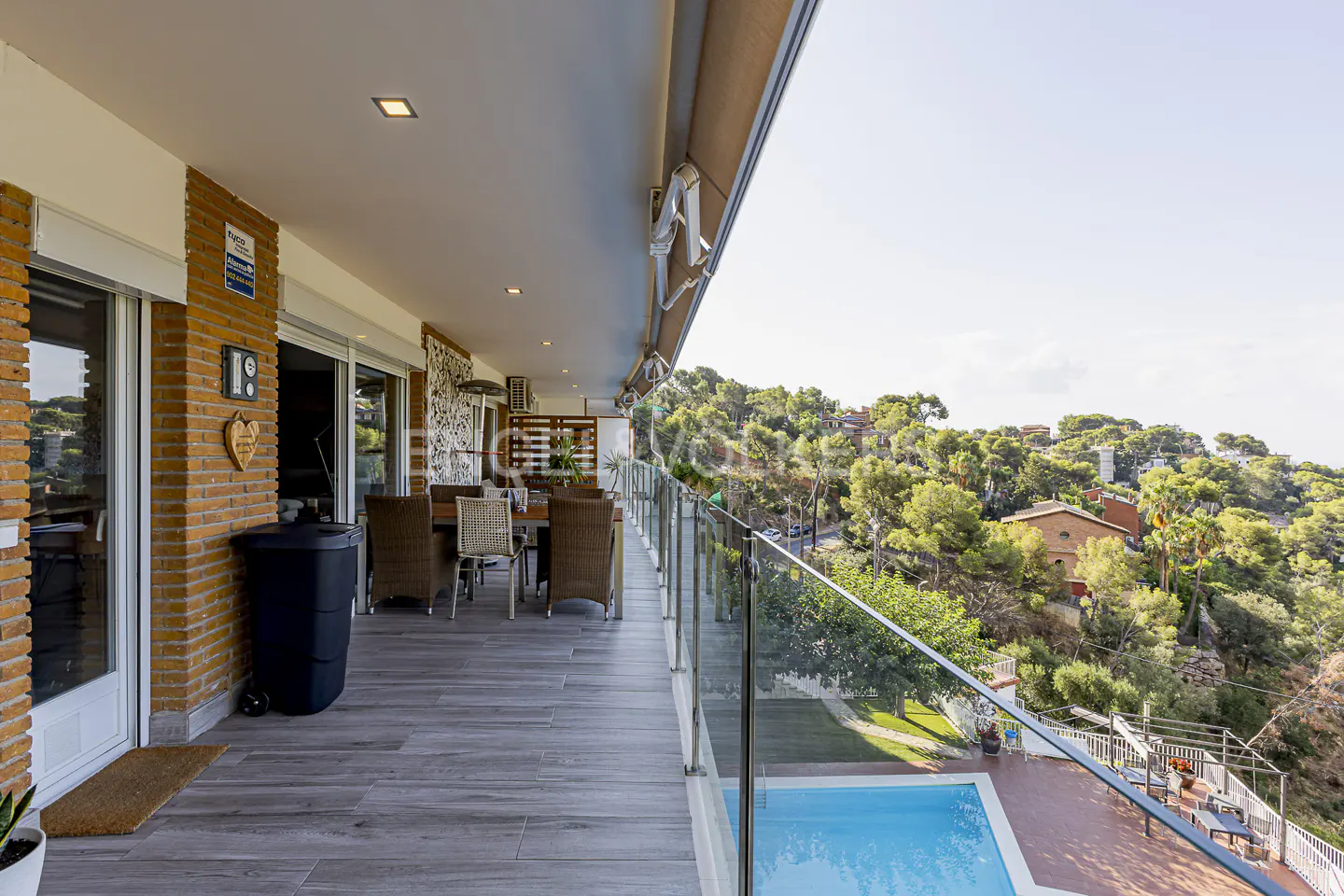 A long balcony with a glass railing overlooks a pool and lush green hillside. Wicker chairs surround a wooden table.