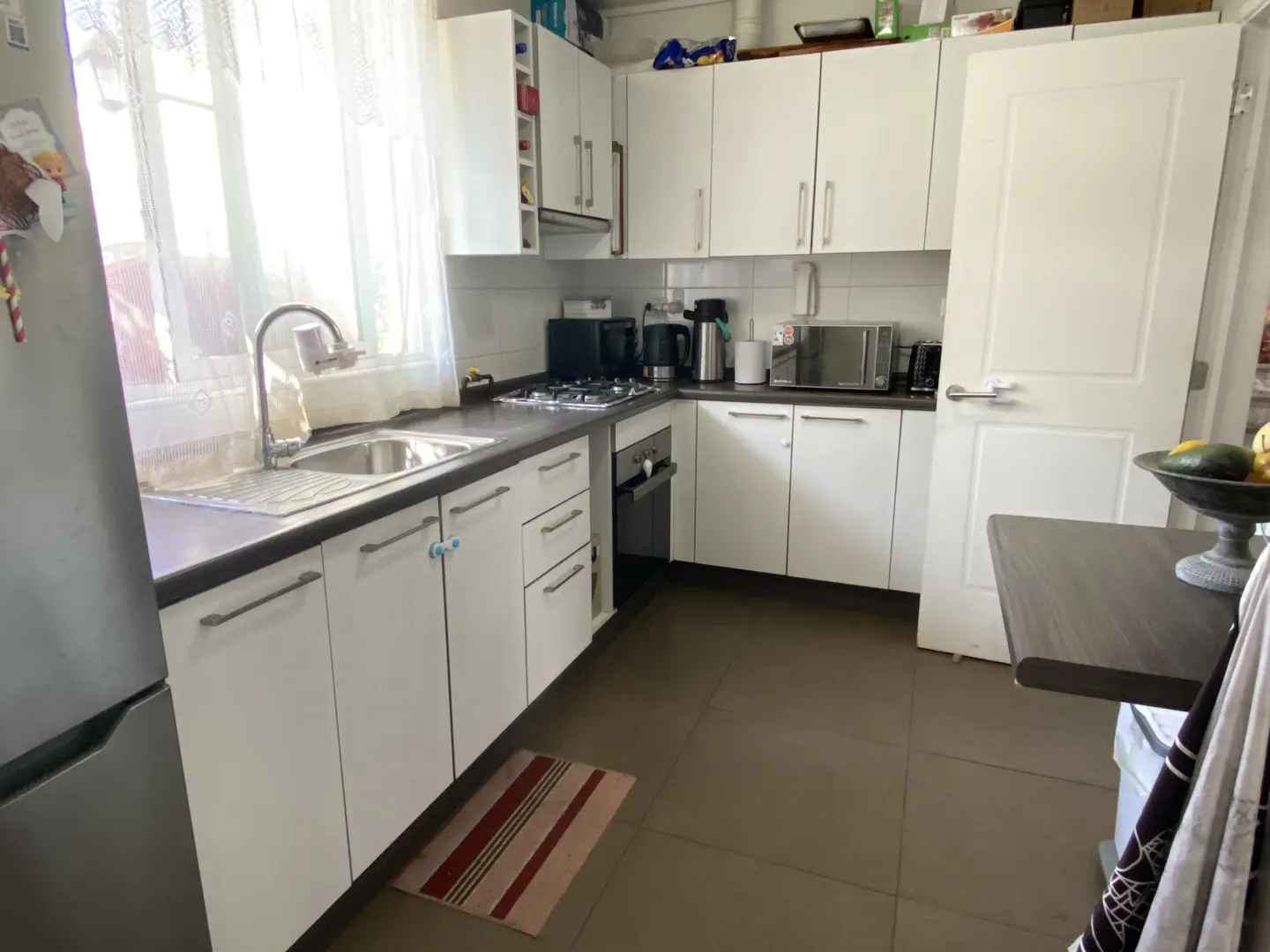 Bright kitchen with white cabinets, dark countertops, and brown tile floor. A stainless steel sink sits under a window with sheer curtains.