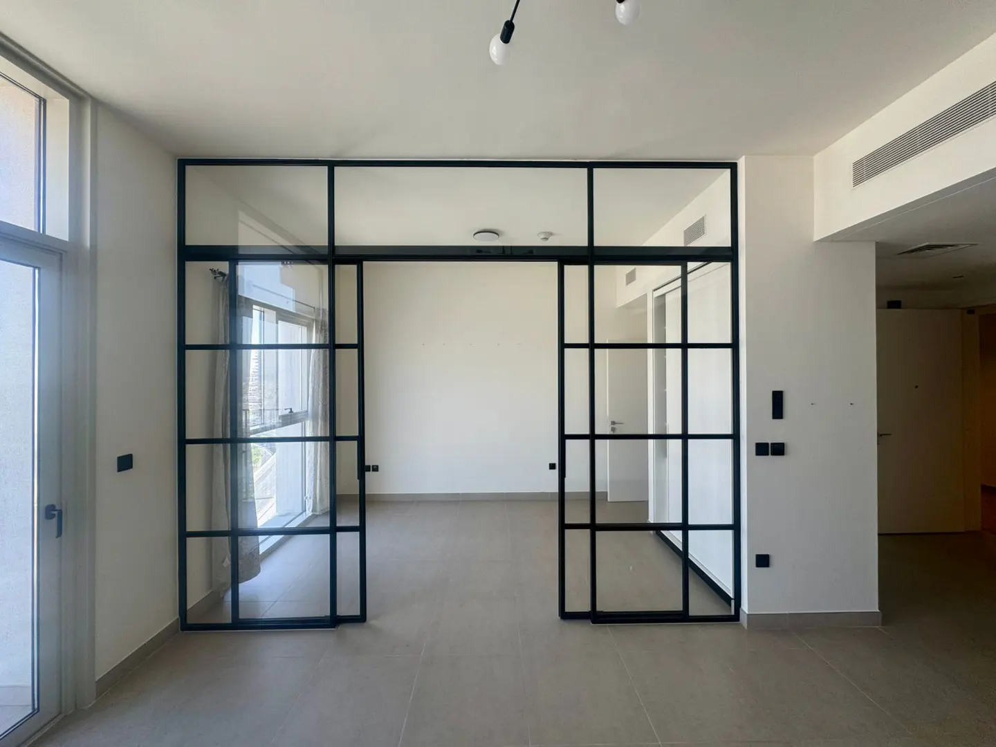 A bright, empty room with sliding black-framed glass doors. The walls are white, and the floor is tiled. A window is visible on the left.